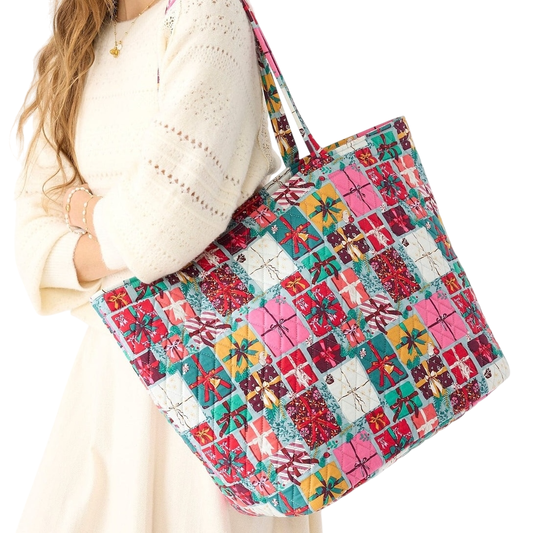 A woman holds a large, colorful patchwork tote bag with a pattern featuring gift box motifs. She is wearing a cream-colored knit dress with delicate patterns.