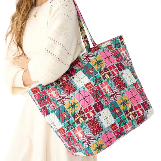 A woman holds a large, colorful patchwork tote bag with a pattern featuring gift box motifs. She is wearing a cream-colored knit dress with delicate patterns.