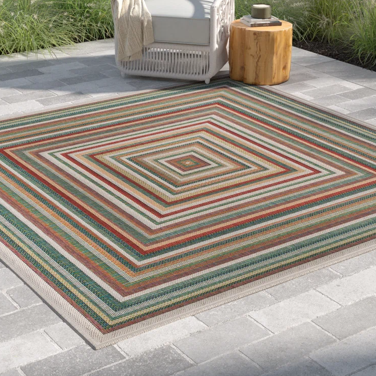 A striped outdoor rug with concentric square patterns in various colors, placed on a tiled patio next to a white chair and a wooden side table.