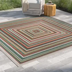 A striped outdoor rug with concentric square patterns in various colors, placed on a tiled patio next to a white chair and a wooden side table.
