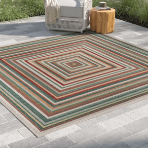 A striped outdoor rug with concentric square patterns in various colors, placed on a tiled patio next to a white chair and a wooden side table.