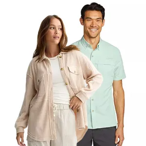 A woman in a beige, button-up, textured overshirt paired with light pants stands next to a man wearing a light green short-sleeve shirt and dark pants.