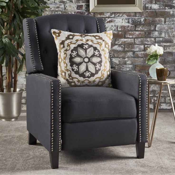 A dark gray recliner with studded rivets trim, featuring a tufted back and square armrests. It is adorned with a decorative pillow with a geometric pattern and set in a modern living space with a brick wall background.