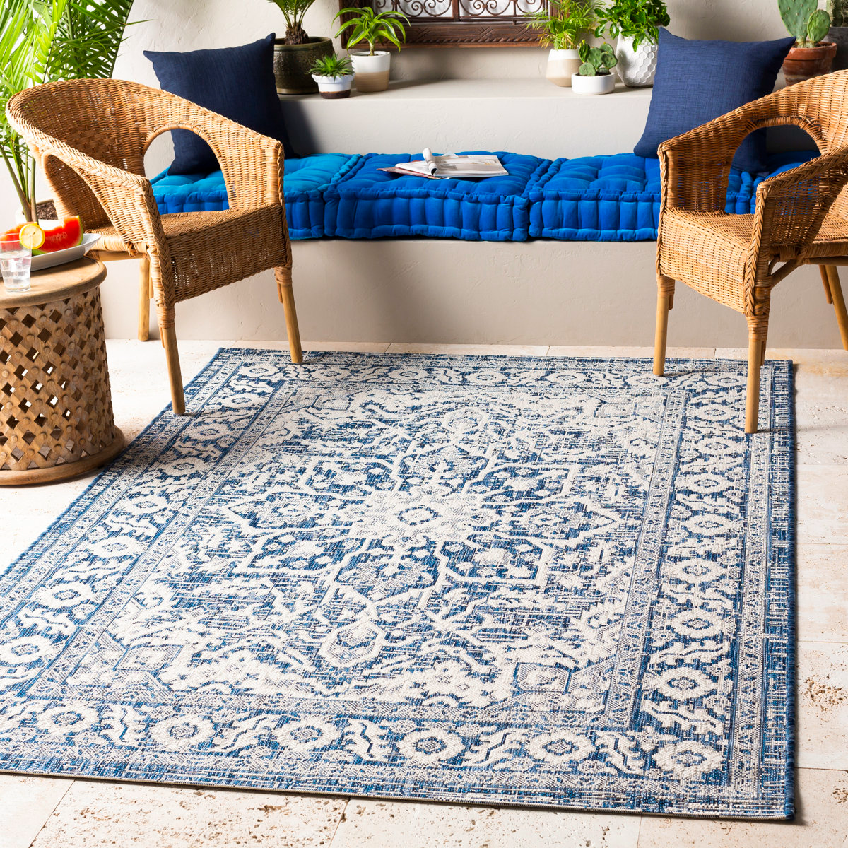 A blue and white area rug with a detailed pattern lies on a tiled floor, surrounded by two wicker chairs and a side table. A cushioned bench with blue pillows and potted plants decorates the background.