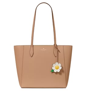 A beige tote bag with two long handles, featuring a floral charm and the \“kate spade new york\“ logo.