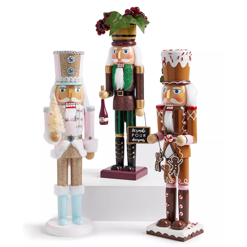 Three decorative nutcrackers: one dressed in pink and gold with a small tree, another in green and purple with a wine bottle, and the third in a gingerbread-themed outfit holding a candy and gingerbread man.