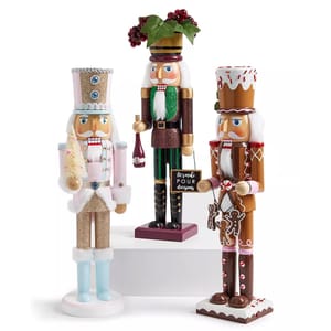 Three decorative nutcrackers: one dressed in pink and gold with a small tree, another in green and purple with a wine bottle, and the third in a gingerbread-themed outfit holding a candy and gingerbread man.