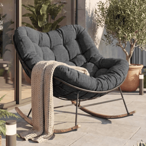 A cushioned rocking chair with a dark fabric and wooden legs is on a patio, accompanied by a knitted beige throw. It's surrounded by potted plants and positioned near large glass doors.