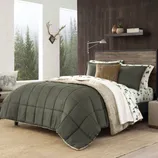 Bedroom with pine green quilted comforter, beige and green tree-patterned sheets, and a decorative bear pillow. The setting features a wooden headboard, wall shelf with plants, and a window with a forest view. A beige rug covers the floor.