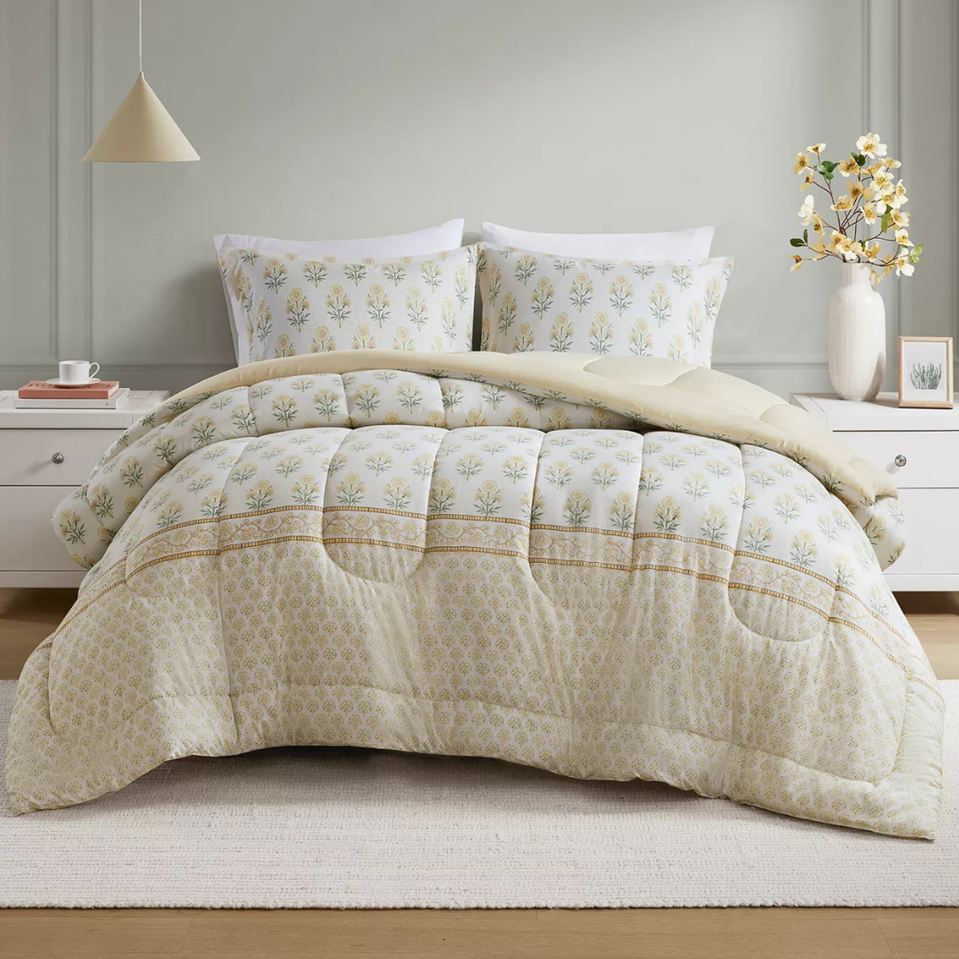 A bed is dressed with a floral-patterned comforter and matching pillowcases. The set is complemented by a simple white nightstand, a neutral pendant light, and a vase with yellow flowers.
