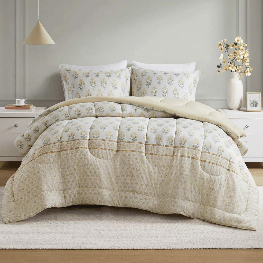 A bed is dressed with a floral-patterned comforter and matching pillowcases. The set is complemented by a simple white nightstand, a neutral pendant light, and a vase with yellow flowers.