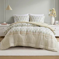 A bed is dressed with a floral-patterned comforter and matching pillowcases. The set is complemented by a simple white nightstand, a neutral pendant light, and a vase with yellow flowers.