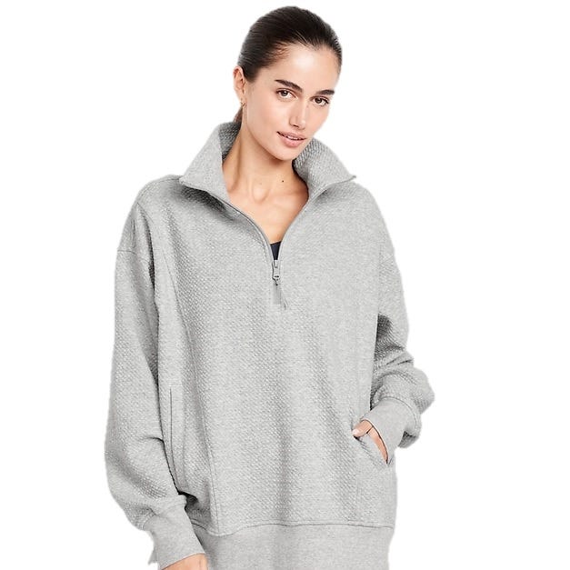 A woman is wearing a light gray, textured pullover with a high collar, partial zip, and long sleeves. The pullover has a loose fit and features pockets.