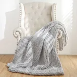 A tufted cream armchair with studded accents holds a chunky, textured light gray knit throw blanket, set on a wooden floor with sunlight streaming through blinds.