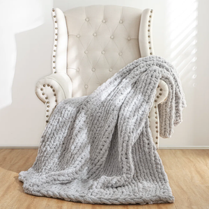A tufted cream armchair with studded accents holds a chunky, textured light gray knit throw blanket, set on a wooden floor with sunlight streaming through blinds.