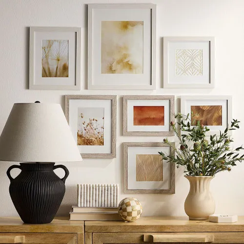 A gallery wall of framed abstract art in neutral tones is displayed above a wooden console table. The table features a black urn-shaped lamp, a beige vase with greenery, stacked books, and a decorative sphere.