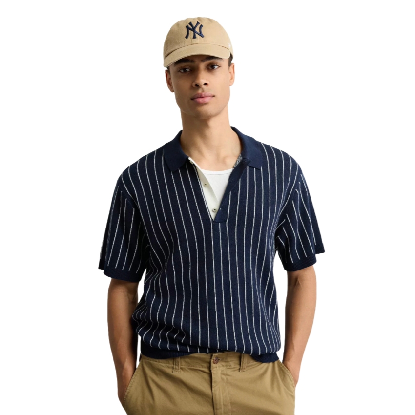 A person wears a navy and white striped polo shirt, a white undershirt, khaki pants, and a beige baseball cap with a black logo featuring \“NY.\“
