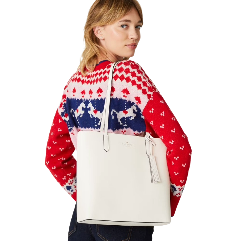 A person wearing a red patterned sweater and dark jeans is holding a white handbag with tassel detail.