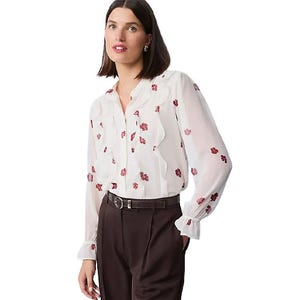 A woman is wearing a white blouse with floral patterns and ruffled detailing, paired with dark high-waisted pants and a belt.