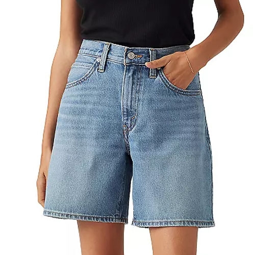 A person is wearing high-waisted, mid-length blue denim shorts with a black top tucked in, one hand in the pocket.