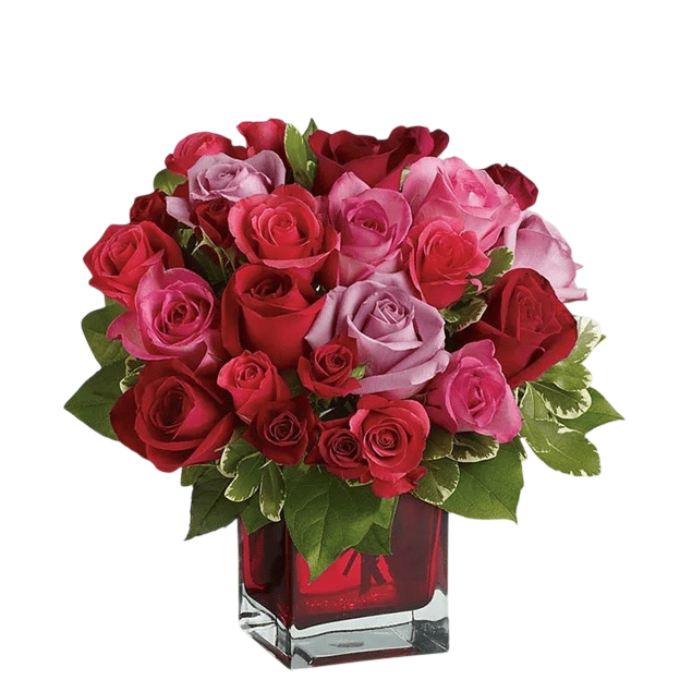 A bouquet of red and pink roses with green leaves arranged in a square glass vase.
