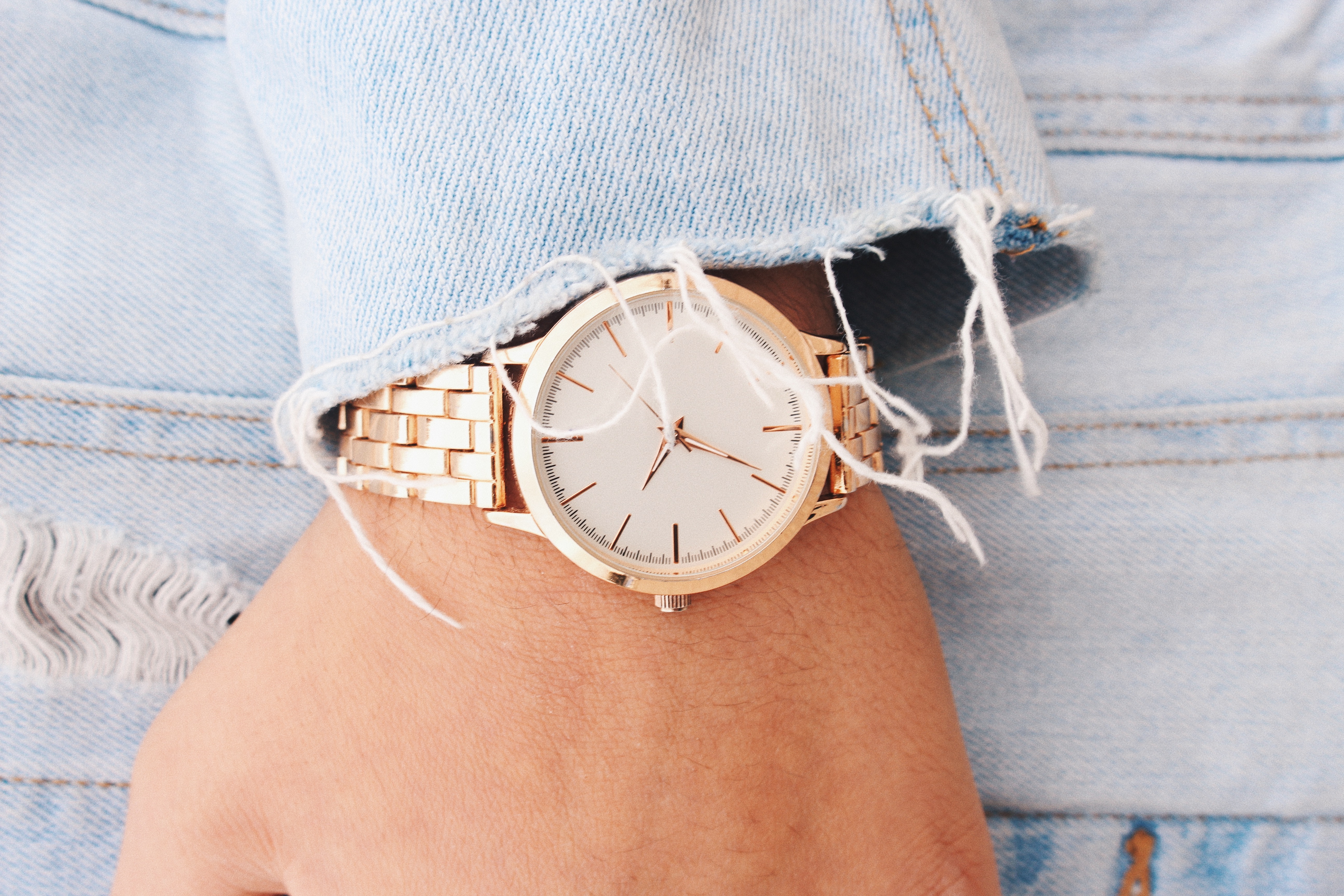 A gold wristwatch with a white dial on a person's wrist, slightly covered by a frayed blue denim sleeve.