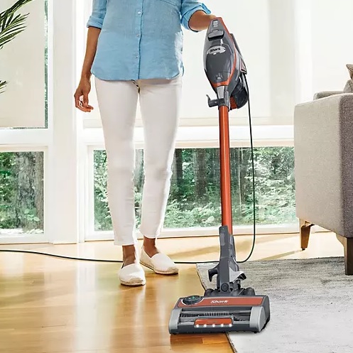 An upright vacuum cleaner being used on a wooden floor in a bright room.