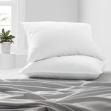 Two white pillows are stacked on a bed covered with a gray sheet. A small potted plant is visible near a window in the background.