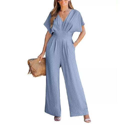 A woman wearing a blue wide leg jumpsuit, white heels, and a tan oversized clutch.