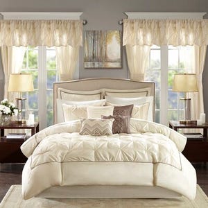 A bedroom set with a cream comforter, decorative pillows, matching curtains, and table lamps on two nightstands, centered around an upholstered bed frame.