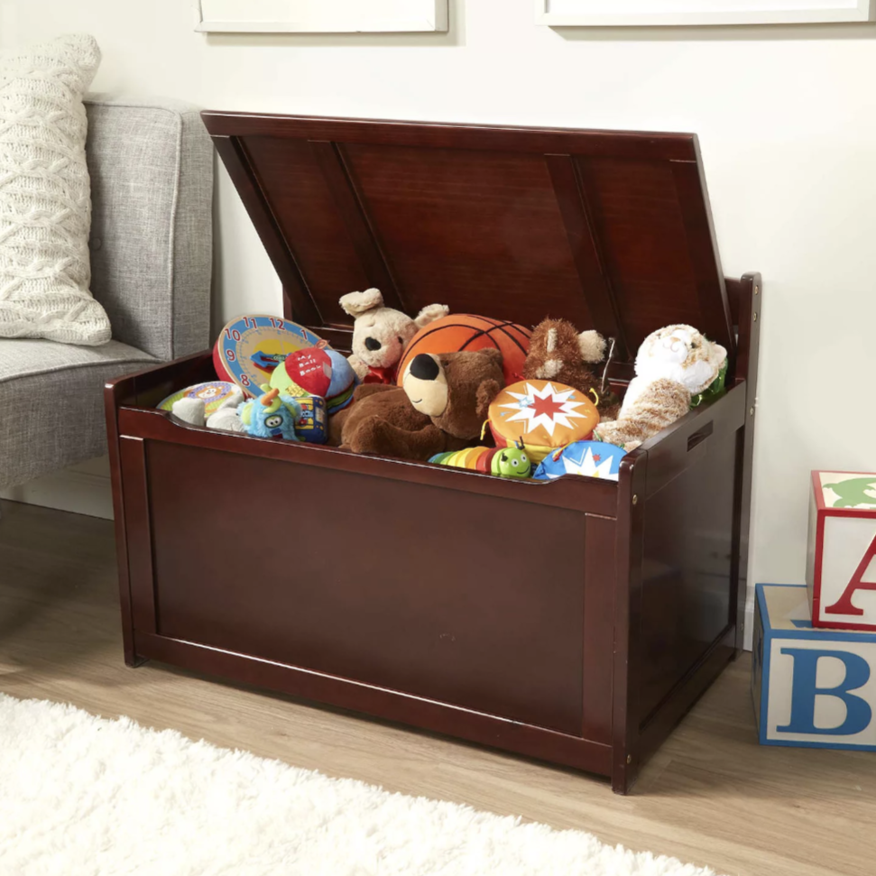 A wooden toy chest filled with various toys, including plush animals, a basketball, and colorful stuffed items. The chest is placed next to a sofa, with alphabet blocks on the floor nearby.