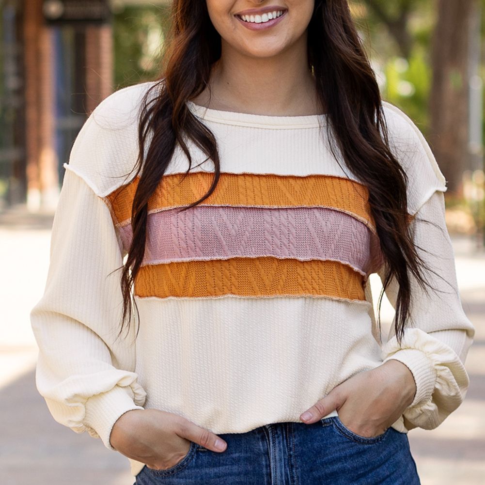 A cream-colored sweater with textured knit bands in rust, pink, and orange across the chest, featuring long sleeves with gathered cuffs and a relaxed fit.
