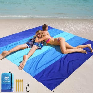 A large, blue-striped beach blanket is laid out on the sand near the water, with two people lounging on it. Included are four yellow stakes, a carabiner, and a compact blue carrying bag labeled \“WIWIGO\“.