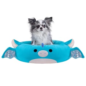 A small dog sits in a plush, blue dragon-shaped pet bed with silver wing details and cartoon-like facial features.