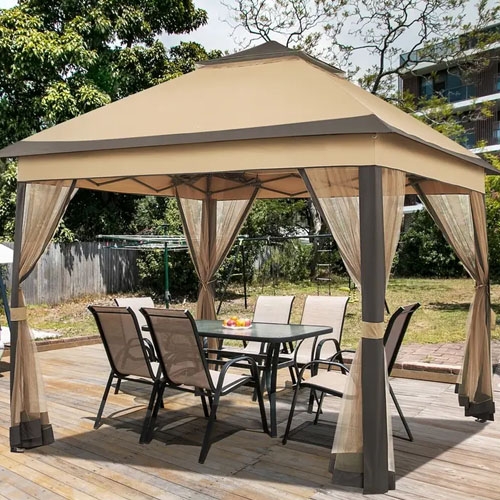 A gazebo with a beige canopy is shown on a wooden deck, featuring mesh curtains and a glass dining table with six chairs underneath.