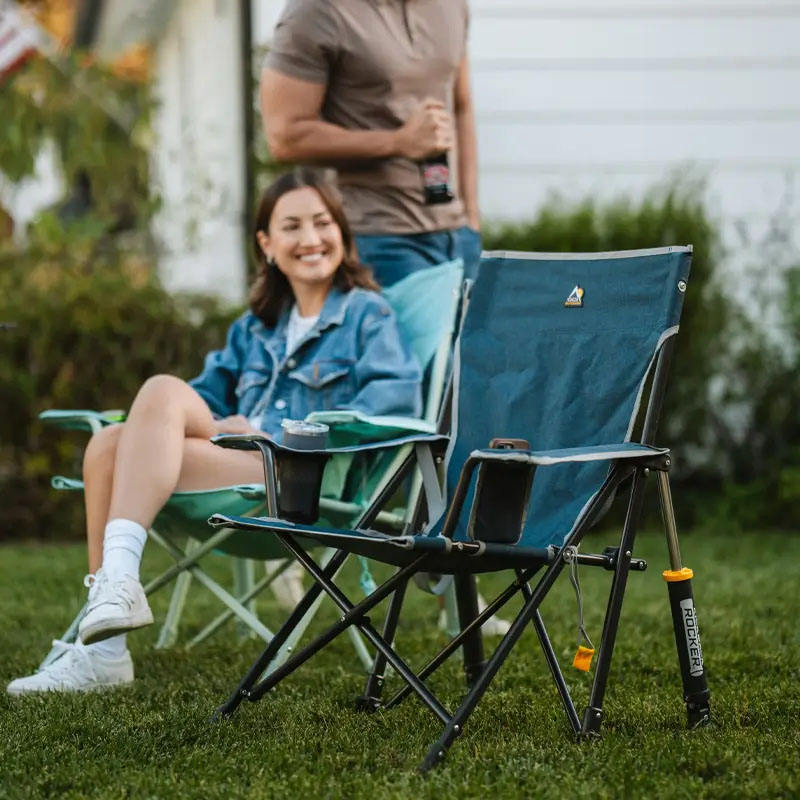 Two people are outdoors; one is sitting in a folding camp chair with a built-in cup holder, and the other is standing nearby holding a beverage. The chair features a rocker mechanism with visible springs.