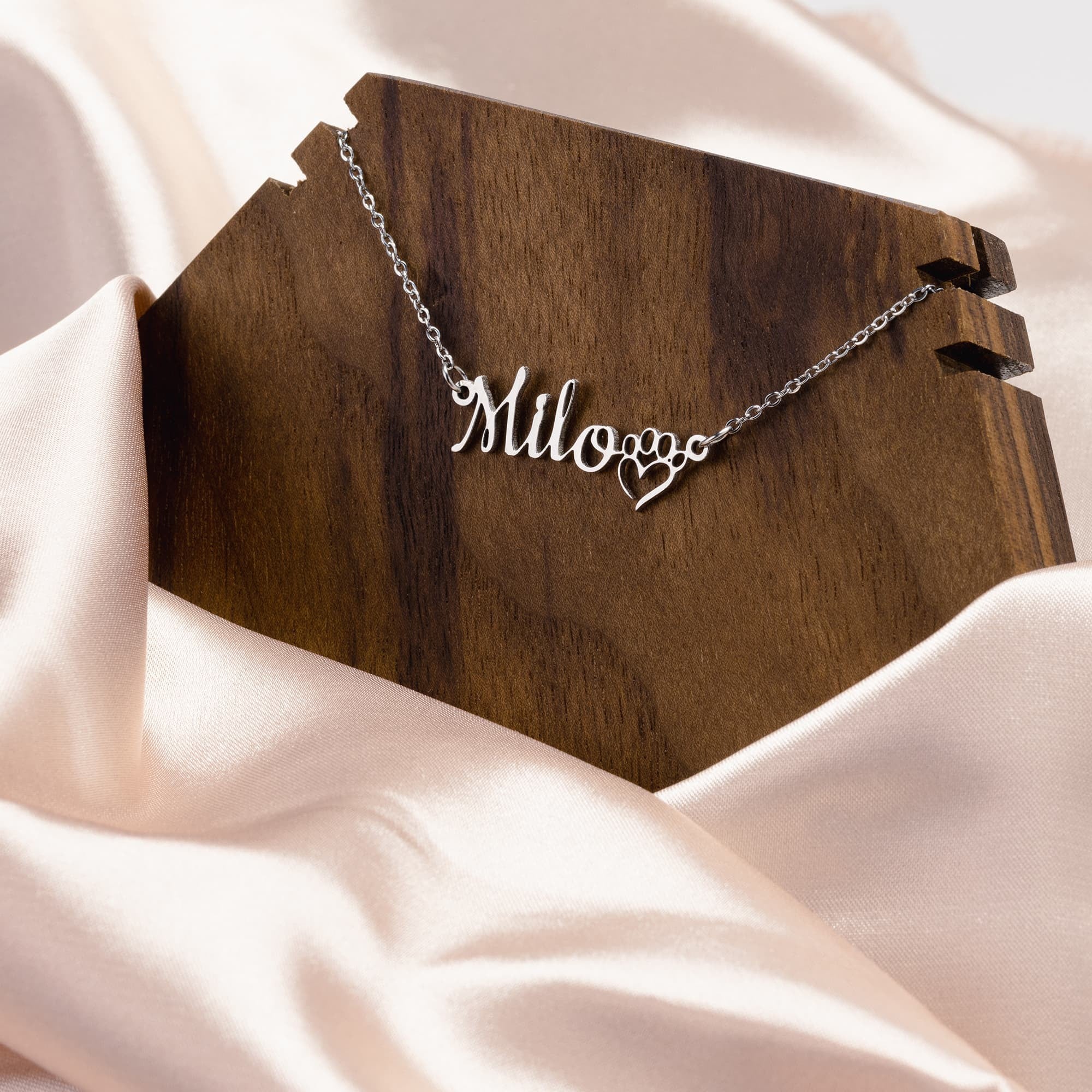 A silver necklace with the word \“Milove\“ and a heart-shaped pendant displayed on a wooden surface with satin fabric backdrop.