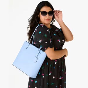 A woman is wearing a floral dress and sunglasses, holding a blue tote bag.