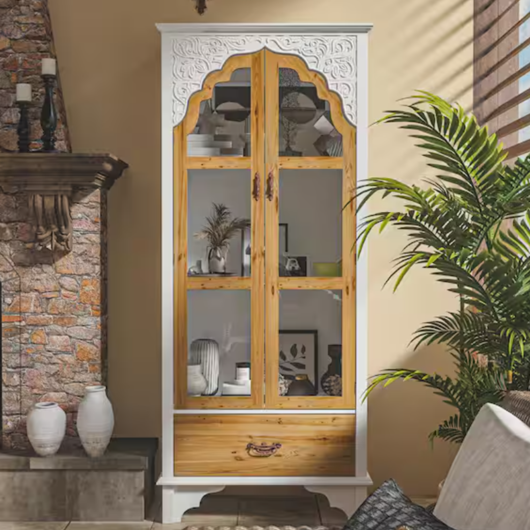 Wooden cabinet with white ornate arch detailing, glass doors, and a bottom drawer. Decor items inside, next to a stone fireplace with candles and near a large indoor plant.