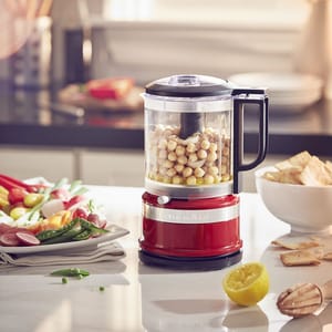 A red KitchenAid Food Chopper with a clear bowl is filled with chickpeas, on a kitchen counter surrounded by various foods.