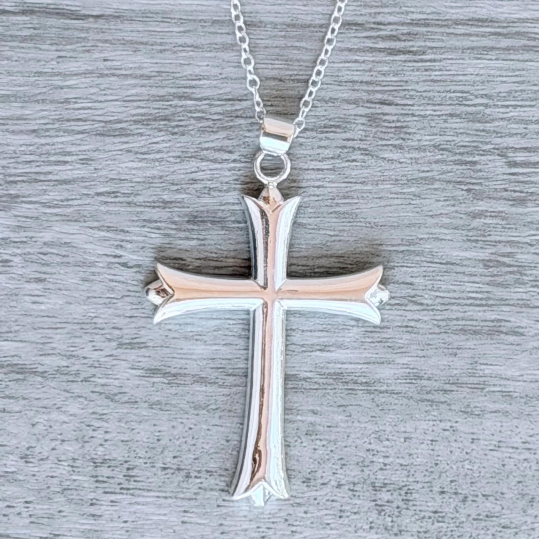 A silver cross pendant with a beveled design hangs on a chain against a textured gray background.
