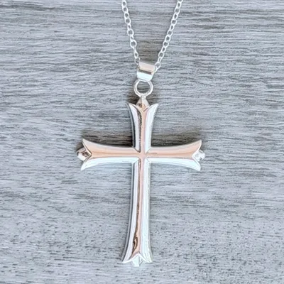 A silver cross pendant with a beveled design hangs on a chain against a textured gray background.