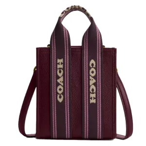 A Coach tote bag with a textured burgundy leather finish and wide straps featuring the Coach logo in white on a pink and black striped background.