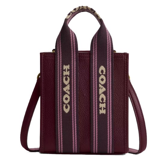 A Coach tote bag with a textured burgundy leather finish and wide straps featuring the Coach logo in white on a pink and black striped background.