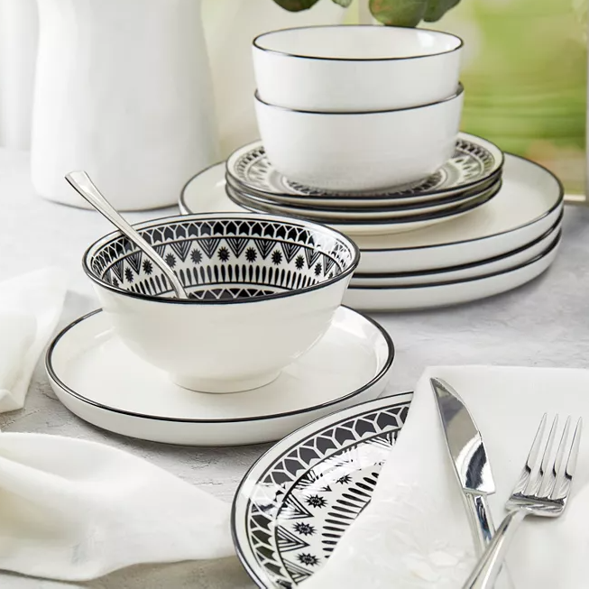 A set of white dinnerware with black geometric patterns, including bowls, plates, and a cup, accompanied by silver cutlery and a white napkin.