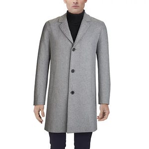 A grey Cole Haan men's topcoat featuring a minimalist design with a three-button front closure, notched lapels, and long sleeves, paired with a black turtleneck sweater and dark trousers.