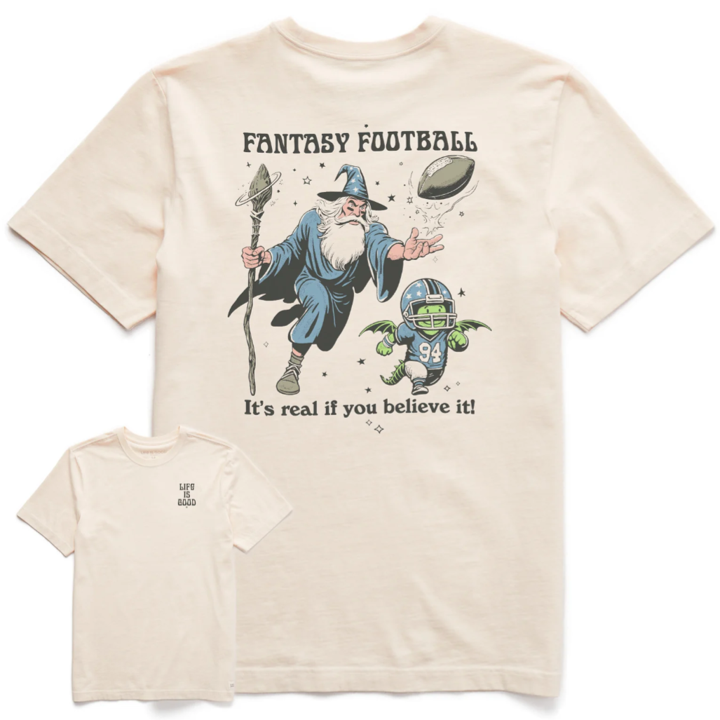 A cream-colored T-shirt featuring a wizard conjuring a flying football with the text \“FANTASY FOOTBALL\“ and \“It's real if you believe it!\“ A small character in football gear with wings is also depicted. The front has \“LIFE IS GOOD\“ printed on the chest.
