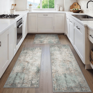 A kitchen features three decorative rugs with a distressed, vintage pattern in muted tones. The rugs are placed on a wooden floor between white cabinetry and appliances.