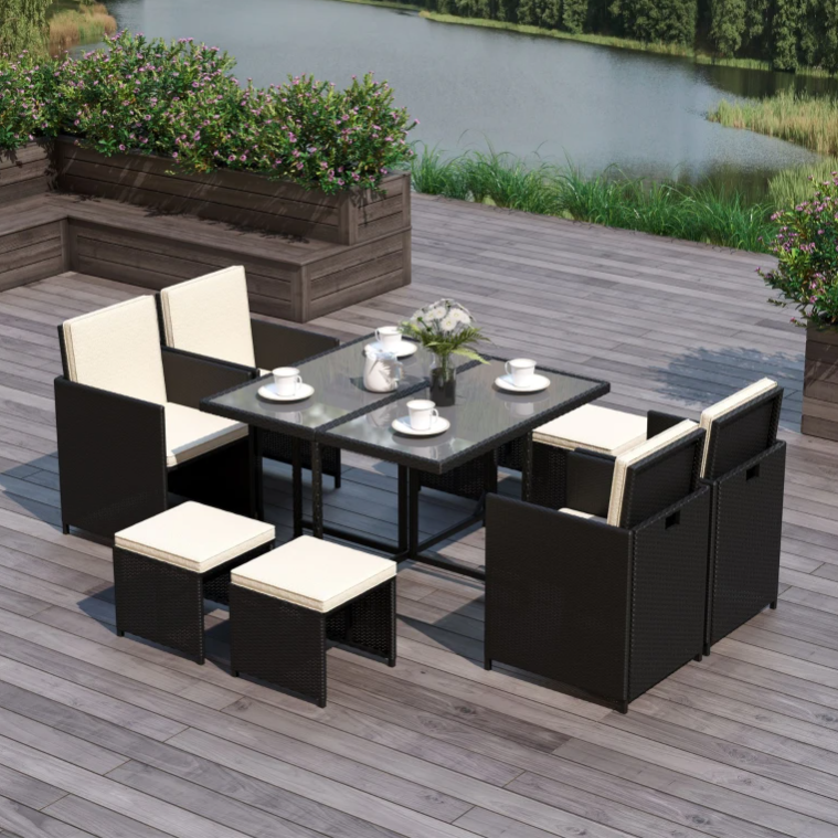 A modern outdoor dining set featuring a glass-top table, four cushioned chairs, and four small cushioned stools, all in a wicker design, situated on a wood deck overlooking a lake.