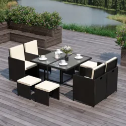 A modern outdoor dining set featuring a glass-top table, four cushioned chairs, and four small cushioned stools, all in a wicker design, situated on a wood deck overlooking a lake.